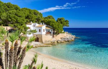 Sonniger Strand der Cala Gat in Cala Ratjada auf Mallorca mit Wohnhaus im Hintergrund.