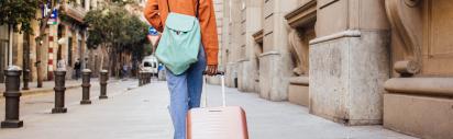 Eine Frau läuft in orangener Jacke mit einem hellblauen Rucksack und einem rötlichen Koffer durch eine Altstadt.