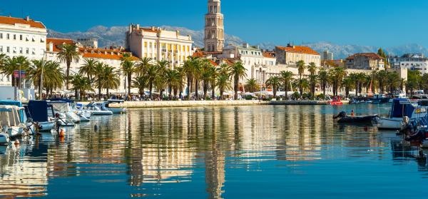 Kathedrale von Split mit Häusern und Palmen drumherum, wie sie sich im Wasser des Hafens spiegelt in welchem Boote stehen.