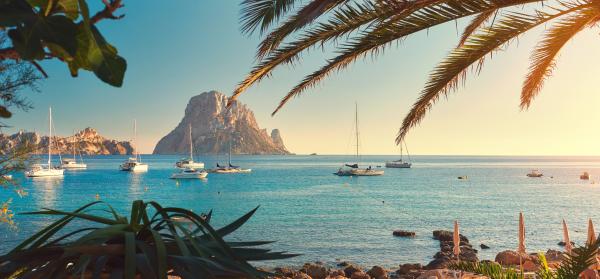 Palmenstrand auf Ibiza mit ankernden Segelbooten im Hintergrund.