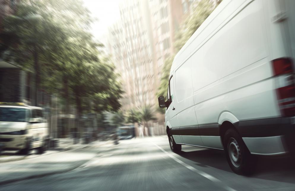 Schnell fahrender Transporter auf einer städtischen Straße, umgeben von Gebäuden und Bäumen im Hintergrund.